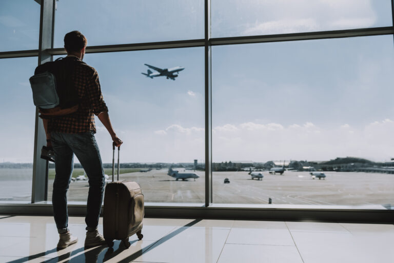 Man with rolling suitcase looking out airport window at airplane landing