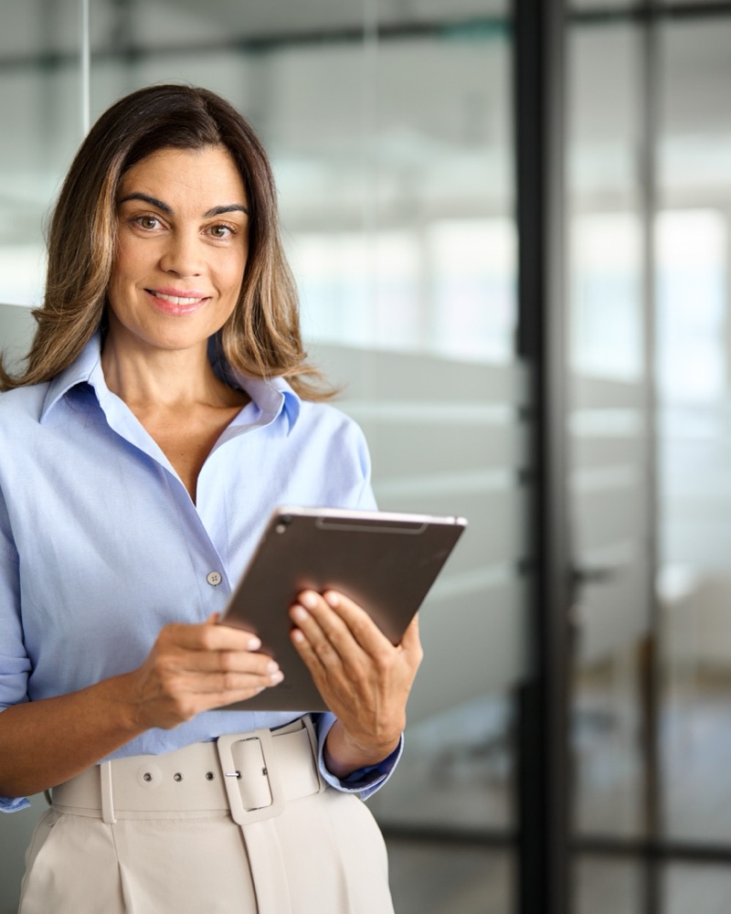 Smiling mature business woman executive holding tablet in office. Portrait. Smiling woman looking at her phone while using laptop to book a trip